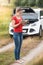 Young woman standing at her broken car in field and reading instructions book