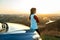 Young woman standing near her car enjoying warm sunset view. Girl traveler leaning on vehicle hood looking at evening horizon