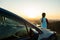 Young woman standing near her car enjoying warm sunset view. Girl traveler leaning on vehicle hood looking at evening horizon