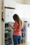 Young woman standing next to an opened refrigerator door, holding a smartphone and ordering fresh fruit and vegetables