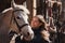 Young woman standing next to white Arabian horse resting in wooden stables box, sun shines on them, closeup detail