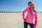 Young woman stands on the beach, wearing a neon pink YOLO shirt with sunglasses, as her hair blows in the wind