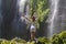 A young woman stands with her back in white clothes at the Sekumpul Great Waterfall in the deep rainforest of Bali Island,