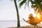 A young woman in straw hat sitting in a hammock swinging between a palm trees on the overseas island sand beach at sunrise time.