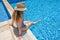 Young woman in straw hat sunbathing sitting on the edge of swimming pool with legs in water