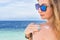 Young woman with sunglasses applyng sun protector cream at her hand on the beach close to tropical turquoise sea under