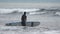 Young woman surfer in wetsuit carrying surfboard walking enter breaking waves to go further into ocean. Slow motion