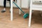 Young woman sweeps laminate flooring in a bright kitchen. The girl sweeps dust and dirt from under the white kitchen table