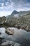 Young woman in swimsuit, warm hat sitting on the rock in the middle of the cold mountain lake, rocky ranges and stones