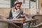 Young woman tourist in sunglasses and hat is sitting in street cafe at table, looking for a way on map, using smartphone