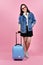 Young woman traveler holding pink suitcase and passport document over pink background.