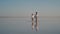 A young woman traveler with a little girl in a shirt is walking on a salt lake, which reflects the sky against a sunset