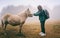 Young woman traveler touching the muzzle of a white pony in a foggy forest, meeting with wildlife