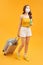 Young woman traveller wearing face mask with suitcase bag on orange background. Trip concept during coronavirus
