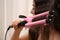 Young woman using modern curling iron against light background, focus on device