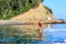 Young woman wakeboarder glides on flat sea surface after motor boat along rocky shoreline at summer sunset. Wakeboarding as outdoo