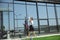 Young woman walking against glass` wall in airport, traveler with small baggage, influencer`s lifestyle