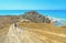 Young woman walks on a deserted dirt road on a promontory towards the sea and lonely beach in summer day