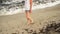 Young woman walks on dirty beach, sand covered with brown washed seaweed, closeup on her legs only