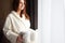 Young woman in warm bathrobe is standing near window and holding basket of rolled up white towels