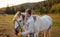 Young woman in warm jacket putting halter on white Arabian horse, blurred meadow and forest background