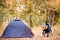 Young woman in warm sweater and black hat resting with guitar near camping tent in wilderness forest