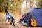 Young woman in warm sweater and black hat resting with guitar near camping tent in wilderness forest