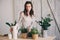 Young woman watering flowerpots at home. Casual lifestyle series in modern scandinavian interior