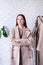 young woman wearing beige pastel coat, standing near clothes rack. Wardrobe change
