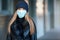 Young woman wearing blue disposable virus face mouth nose mask and warm beanie hat, blurred building wall background. Coronavirus