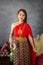 young woman wearing a original traditional red Thai dress and golden ornaments stands holding a jasmine garland. Thai national