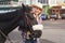 Young woman wearing shirt, gloves and straw hat standing behind dark brown horse, smiling, blurred city street with parked cars in