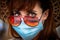 A young woman wearing a surgical mask, red sunglasses and a hat ready for holidays after coronavirus lock down
