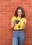Young woman wearing yellow shirt, holding camera and showing that everything is OK against brick wall