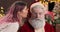 A young woman whispers something in the ear of a surprised Santa Claus sitting in a New Year\'s studio with lights, next