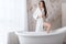 Young woman in a white bathrobe sitting at the side of freestanding white bath