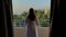 Young woman in a white coat opening curtains and enjoying sea view from terrace in morning.