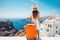 Young woman in white dress and hat with orange suitcase on Santorini island, Greece, Happy moment with young woman rear view