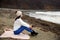 Young woman in white sweater, hat with pom-pom, sitting on the shore of the Japanese sea with closed eyes, inhaling the aroma of w