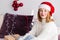 A young woman in a white sweater, Santa Claus hat, with New Year gifts sits on the sofa, looks at the camera and smiles