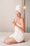 Young woman in white towel apply moisturizer cream in bedroom. Skin care, spa concept