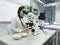 A young woman in a white uniform is working in a laboratory behind a microscope .