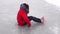 Young woman in winter clothes and high-heeled shoes sits on ice after fall. Crazy female crawling on outdoor ice rink in