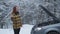 A young woman in a winter forest is standing on the road with a broken car, stopping cars, waiting for help