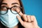 Young woman wiping foggy glasses caused by wearing disposable mask on blue background, closeup. Protective measure during