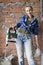 Young woman in work clothes and a protective helmet stands next to a brick wall and holds a heavy hammer drill