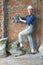 Young woman in work clothes and a protective helmet stands next to a brick wall and makes a hole with a heavy hammer drill