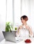 Young woman working at home with notebook sitting on near window drinking tea from the cup and eating cherry