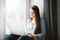 Young woman works at laptop and sits at windowsill