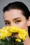 young woman with yellow eyeshadows covering face with blooming flowers isolated on grey.